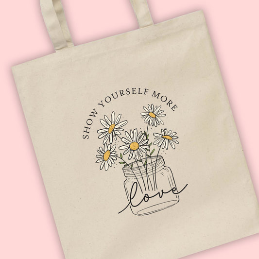A natural tote bag with ‘Show yourself More Love’ written with an illustration of a vase of daisy flowers